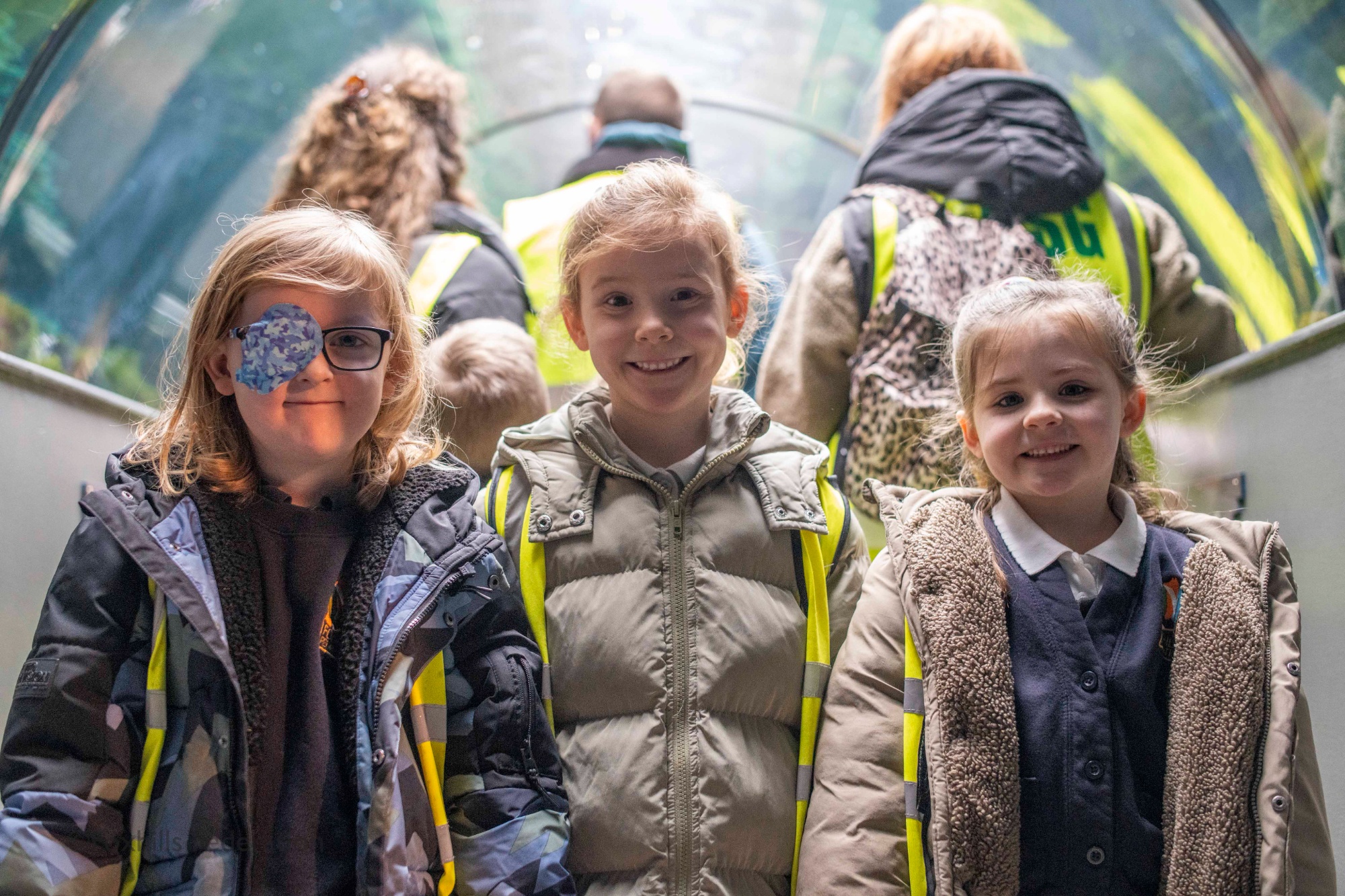 Three children smile at the camera in a glass tunnel.